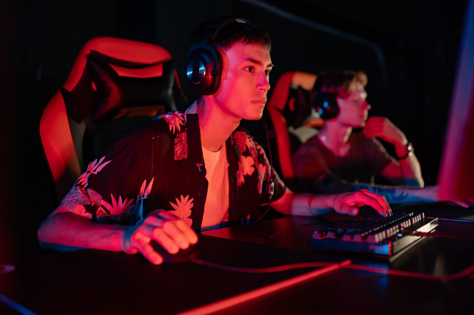 Two players in headphones side by side at a desk in red neon light, focused on their screens
