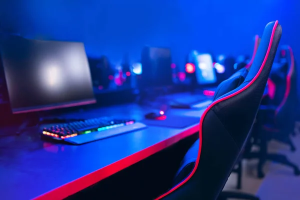 An empty gaming station under blue and red neon: curved monitor, RGB keyboard and a black racing chair